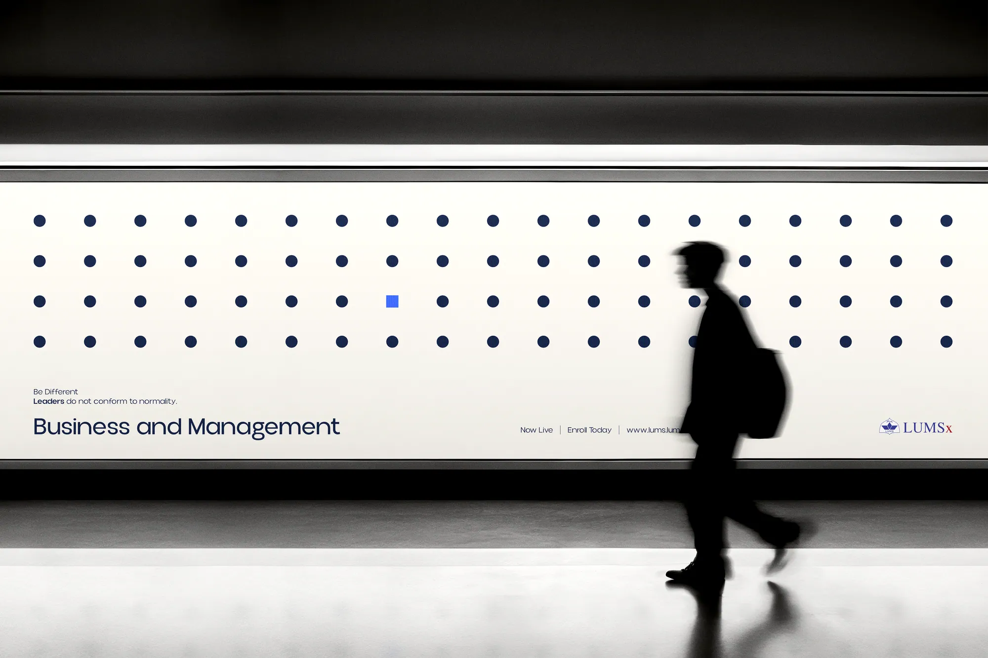 A LUMSx Business and Management billboard on a station platform, with a dotted pattern and a commuter walking past