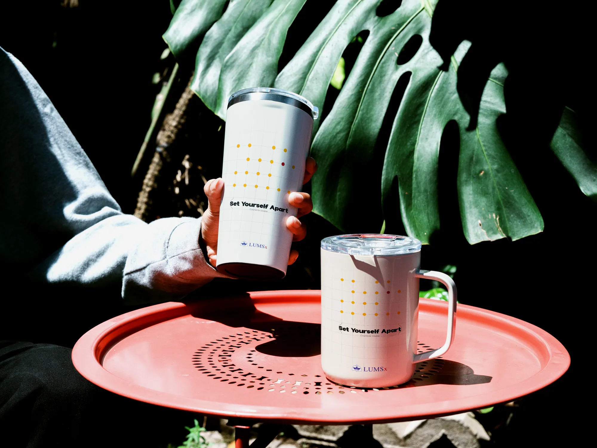 A white LUMSx tumbler and mug with a dotted pattern on a coral tray under palm leaves