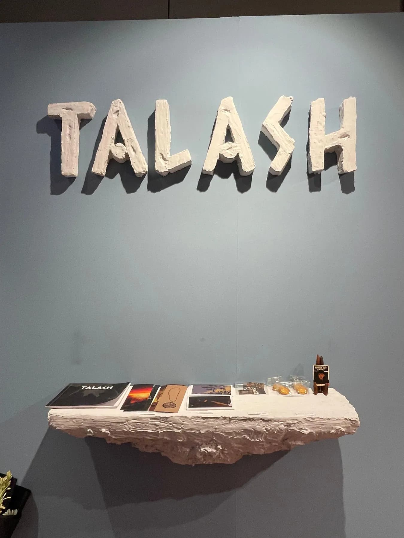 The Talaash title in raised letters on a pale wall above a shelf of prints